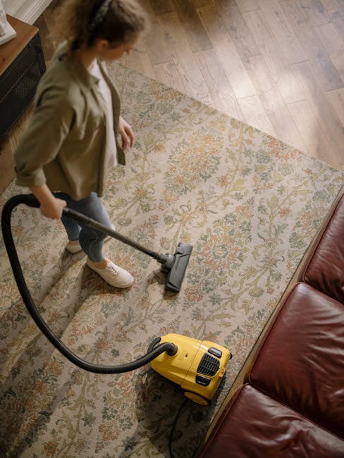 A woman using a yellow vacuum cleaner to deep clean a patterned area rug in a living room. The room features wooden flooring and a red leather sofa. The woman is dressed casually in a beige jacket and