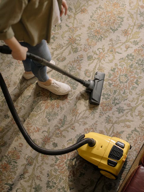 A woman using a yellow vacuum cleaner to deep clean a patterned area rug in a living room. The room features wooden flooring and a red leather sofa. The woman is dressed casually in a beige jacket and