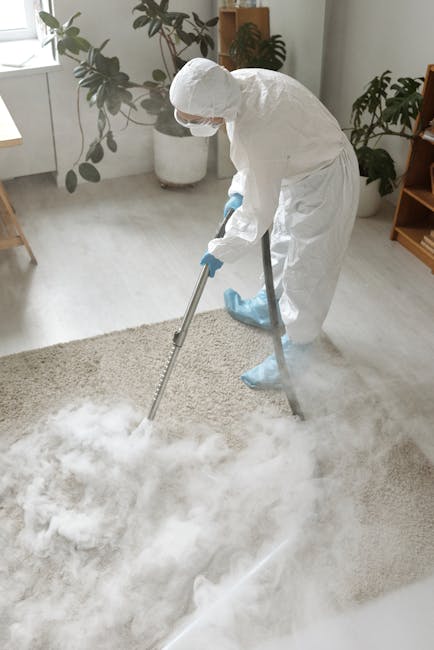 A professional cleaner dressed in a full white protective suit, mask, gloves, and shoe covers is performing surface cleaning on a plush beige carpet in a well-lit living room. The cleaner is using a v
